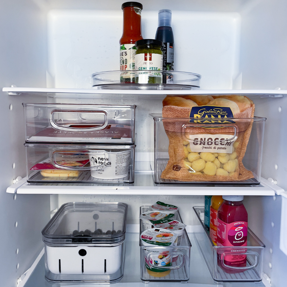 Ein gut organisierter Kühlschrank enthält Getränke, Soßen, Käse, Eier, Obst, Joghurt, Saft und Kartoffelschalen, die alle in durchsichtigen Behältern und auf Regalen gelagert sind.