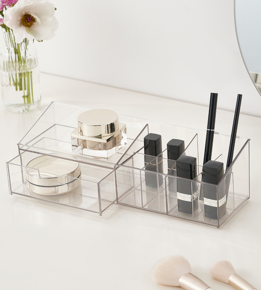 Der transparente Multi-Organizer KLAR (30.6x9x9cm) von BNB x VCN ordnet goldene Döschen, schwarze Lippenstifte und Make-up-Pinsel auf dem weißen Schminktisch neben Vase und rundem Spiegel.