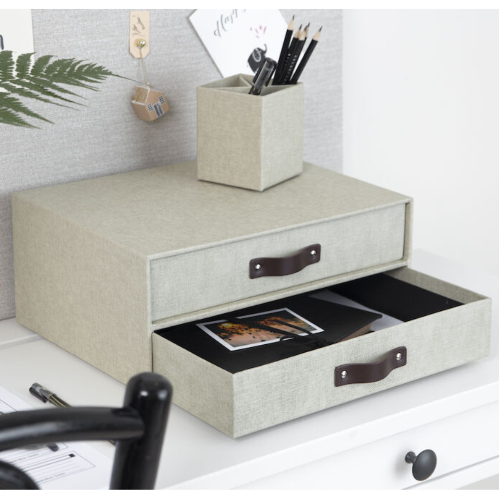 Eine Bigso BIRGER Schubladenbox 2 Fächer in Leinen Canvas steht auf einem weißen Schreibtisch. Die untere Schublade ist geöffnet und zeigt Fotos im Inneren. Oben befindet sich ein passender Stifthalter mit Stiften und Bleistiften.