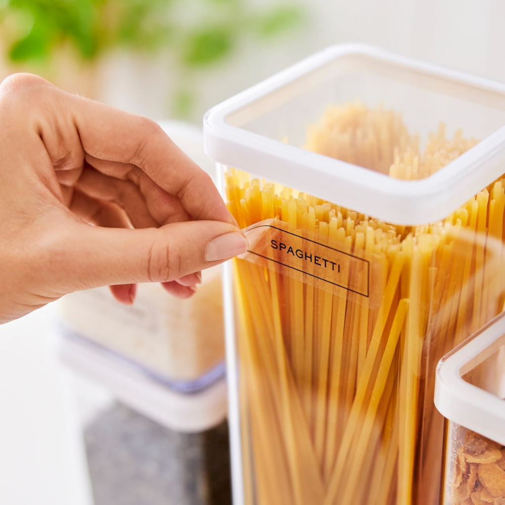 Eine Hand klebt ein Etikett der Rotho LOFT Aufkleber - Küchenorganisation mit der Aufschrift SPAGHETTI auf einen durchsichtigen Behälter mit ungekochten Spaghetti, während im Hintergrund weitere beschriftete Lebensmittelbehälter aus derselben Serie zu sehen sind.