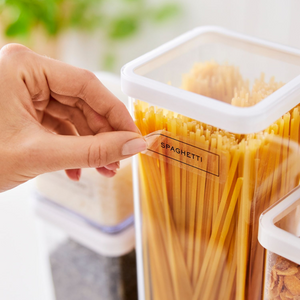 Eine Hand klebt ein Etikett der Rotho LOFT Aufkleber - Küchenorganisation mit der Aufschrift SPAGHETTI auf einen durchsichtigen Behälter mit ungekochten Spaghetti, während im Hintergrund weitere beschriftete Lebensmittelbehälter aus derselben Serie zu sehen sind.