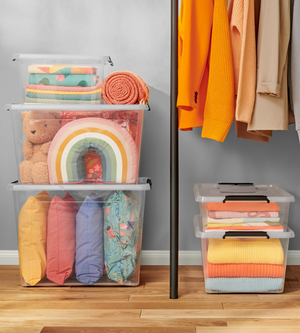 Die Orplast SimpleStore Box WHEEL (40L, 58x39x25 cm) hält bunte Kissen, einen Teddybär, ein Regenbogenkissen und Decken. Daneben hängen gelbe und orange Kleidung. Die stapelbaren Boxen stehen ordentlich auf dem Holzfußboden.
