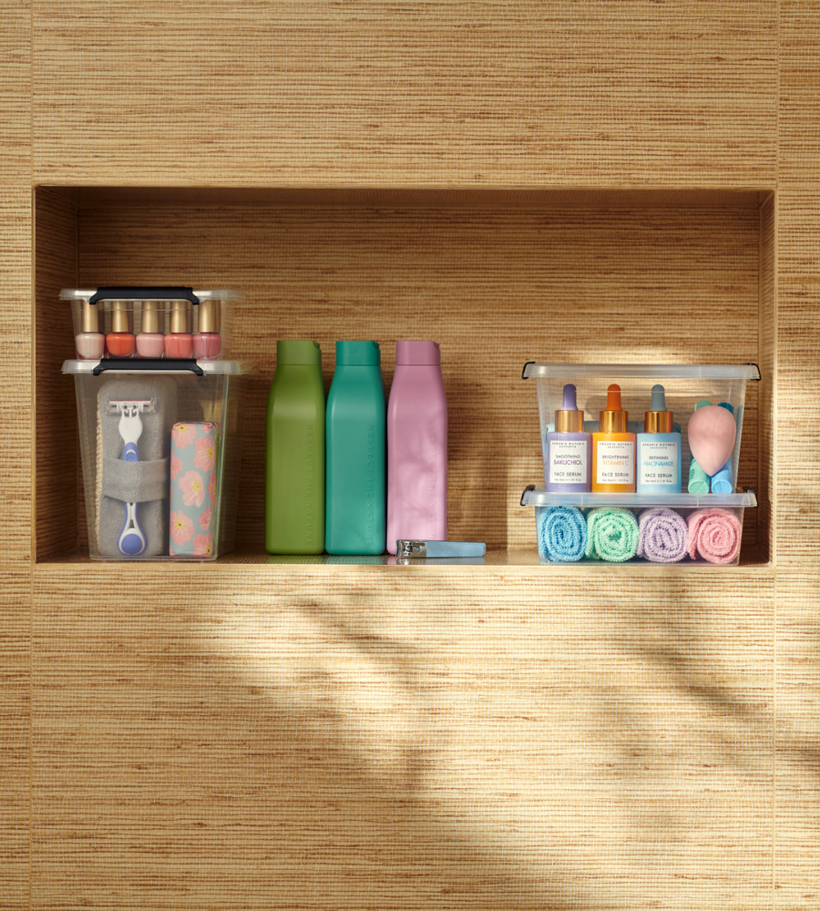 Auf drei Badezimmerregalen werden die Toilettenartikel in der Orplast SimpleStore Box - 29x20x12 cm (4,5 L) ordentlich aufbewahrt: Nagellack, Rasierer, Hautpflegeserum, Make-up-Schwämme, Waschlappen und Flaschen vor einer gewebten Wand.