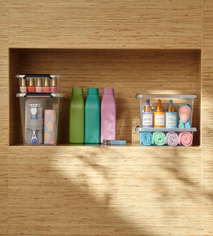 Auf drei Badezimmerregalen werden die Toilettenartikel in der Orplast SimpleStore Box - 29x20x12 cm (4,5 L) ordentlich aufbewahrt: Nagellack, Rasierer, Hautpflegeserum, Make-up-Schwämme, Waschlappen und Flaschen vor einer gewebten Wand.