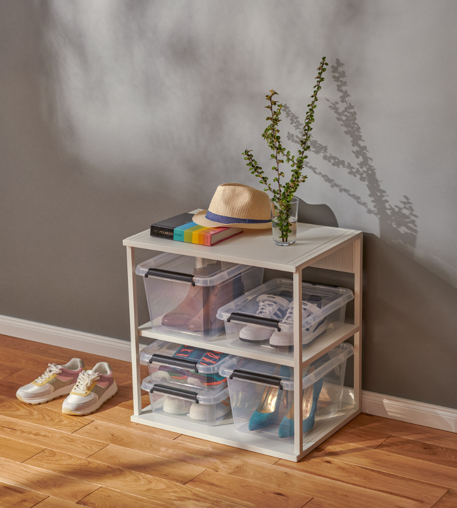 Ein weißes Regal enthält Orplast SimpleStore Box - 39x29x11 cm (8,5 L) Aufbewahrungsboxen mit Schuhen darin. Obenauf stehen eine Topfpflanze, Bücher und ein Strohhut. Zwei Paar Turnschuhe stehen auf dem Holzboden vor einer grauen Wand.