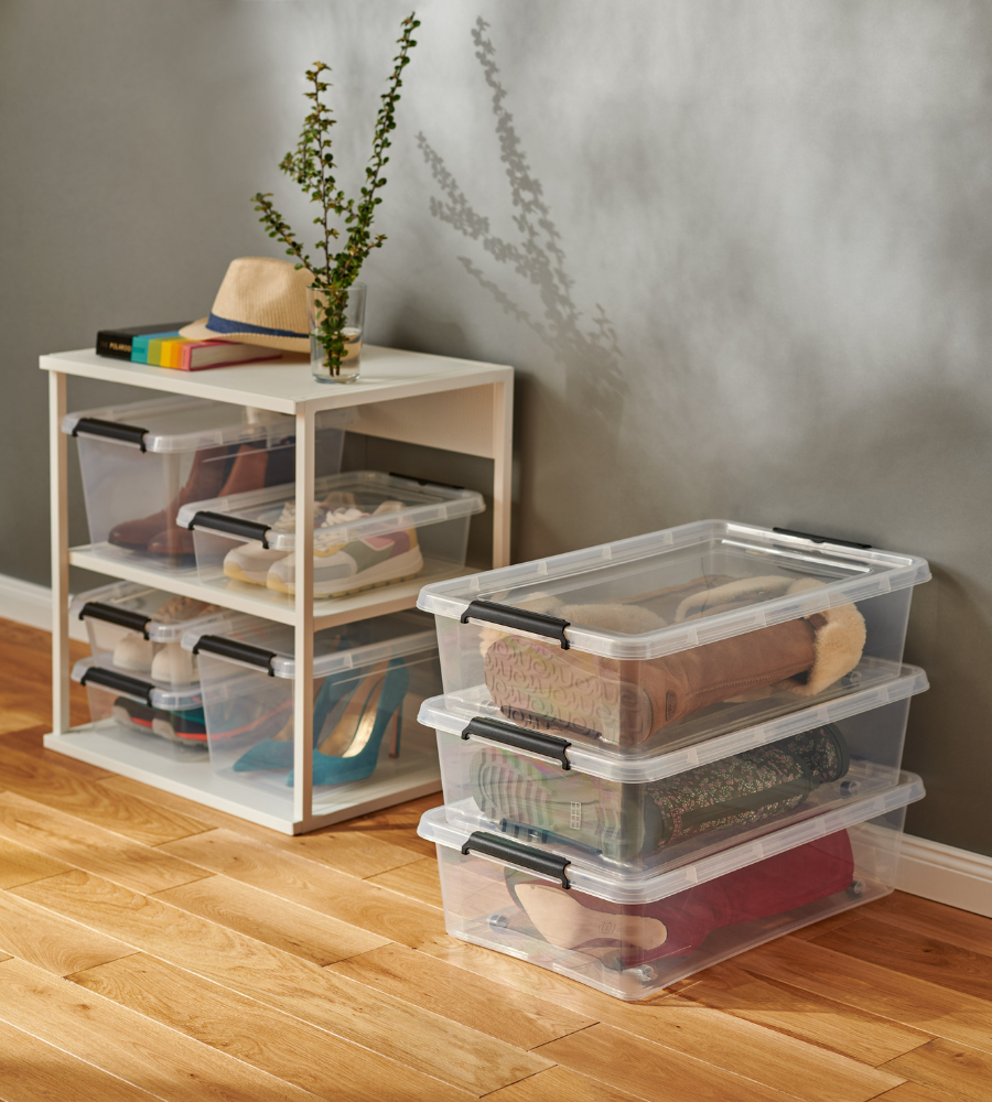 Drei BPA-freie Orplast SimpleStore Boxen (39x29x16,5 cm, 12,5L) sind voll mit Schuhen auf einen Holzfußboden gestapelt, neben einem kleinen weißen Regal mit weiteren klaren Boxen, Büchern, Hut und Vase mit Grünpflanze.