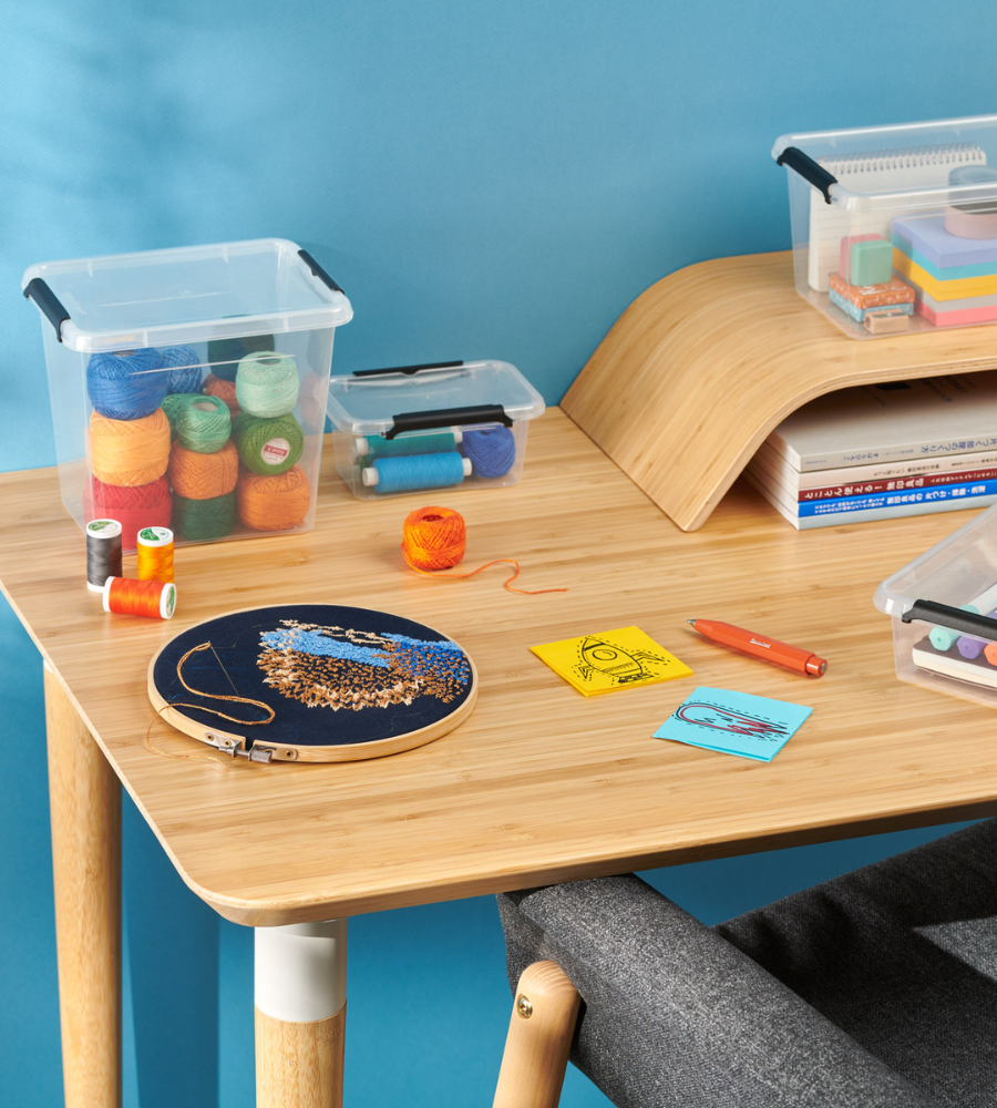 Ein hölzerner Schreibtisch mit bunten Stickgarnen, der Orplast SimpleStore Box STACK&NEST (39x39x26cm, 25L), Büchern, einem Stickrahmen in Arbeit, skizzenhaften Haftnotizen und einem Stift auf blauem Hintergrund. Ein grauer Stuhl ist teilweise sichtbar.