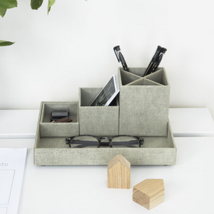 Der LENA Schreibtisch Organizer - Linnen Canvas von Bigso in Grau bietet Platz für Stifte, ein Telefon und mehr. Holzblöcke und Papier liegen auf dem weißen Schreibtisch, während eine grüne Pflanze aus der Ecke lugt - ideal für Schreibtisch Ordnung.