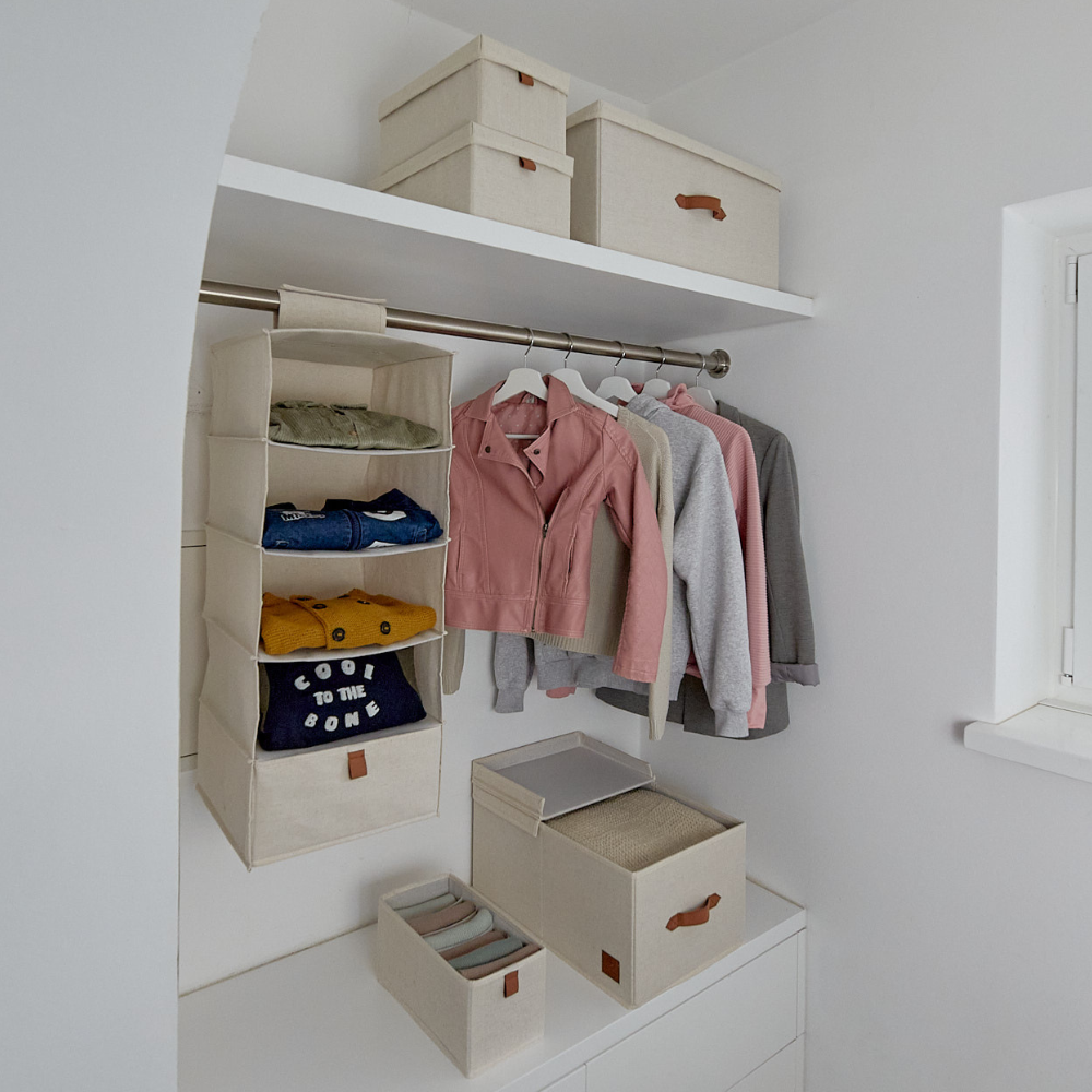 Ein aufgeräumter Kleiderschrank mit Love it Store it PREMIUM Aufbewahrungsboxen mit Deckel Beige (Set 5 Stk.) mit verstärkten Kartonwänden, hängenden Stoffböden für gefaltete Kleidung, sowie einer Stange mit Jacken und Hemden. Ein Fenster auf der rechten Seite bringt Ordnung ins Zuhause.