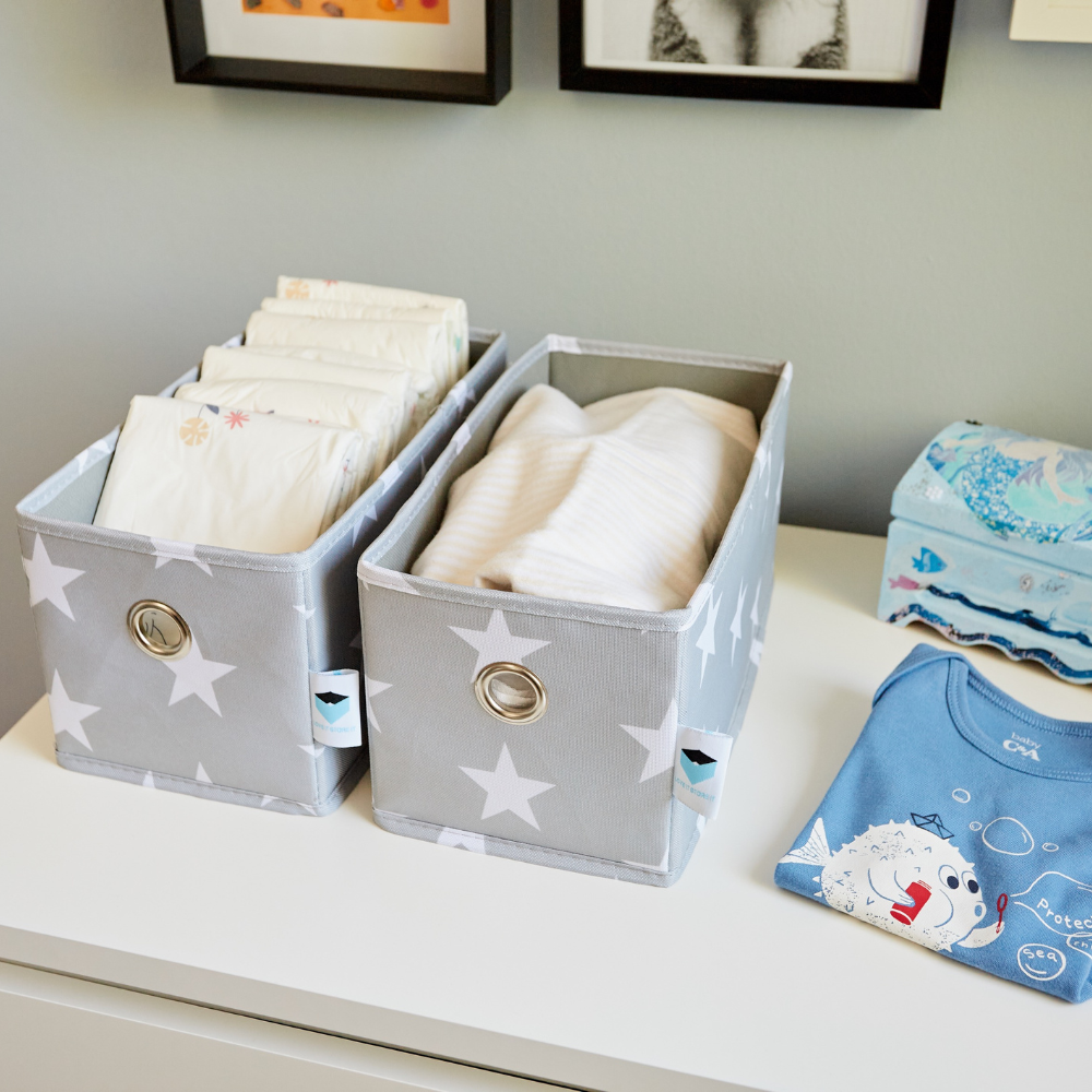 Two STAR Organizer schmal Grau - Set 2 Stk von Love it Store it, mit weißen Sternen, halten Babywindeln und Kleidung auf einer Kommode. Auch auf der Kommode befinden sich eine blaue Taschentuchbox und ein gefalteter blauer Onesie, wodurch ein praktischer Organizer unter der Wandkunst entsteht.