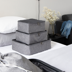 Drei Bigso TRAVEL Organizer S Würfel in Grau liegen gestapelt auf einem ordentlich gemachten Bett. Auf dem Nachttisch liegen Trockenblumen, Brillen, Jeans und Teile eines Kissens und Koffers - ideal für alle, die Koffer organisieren wollen.