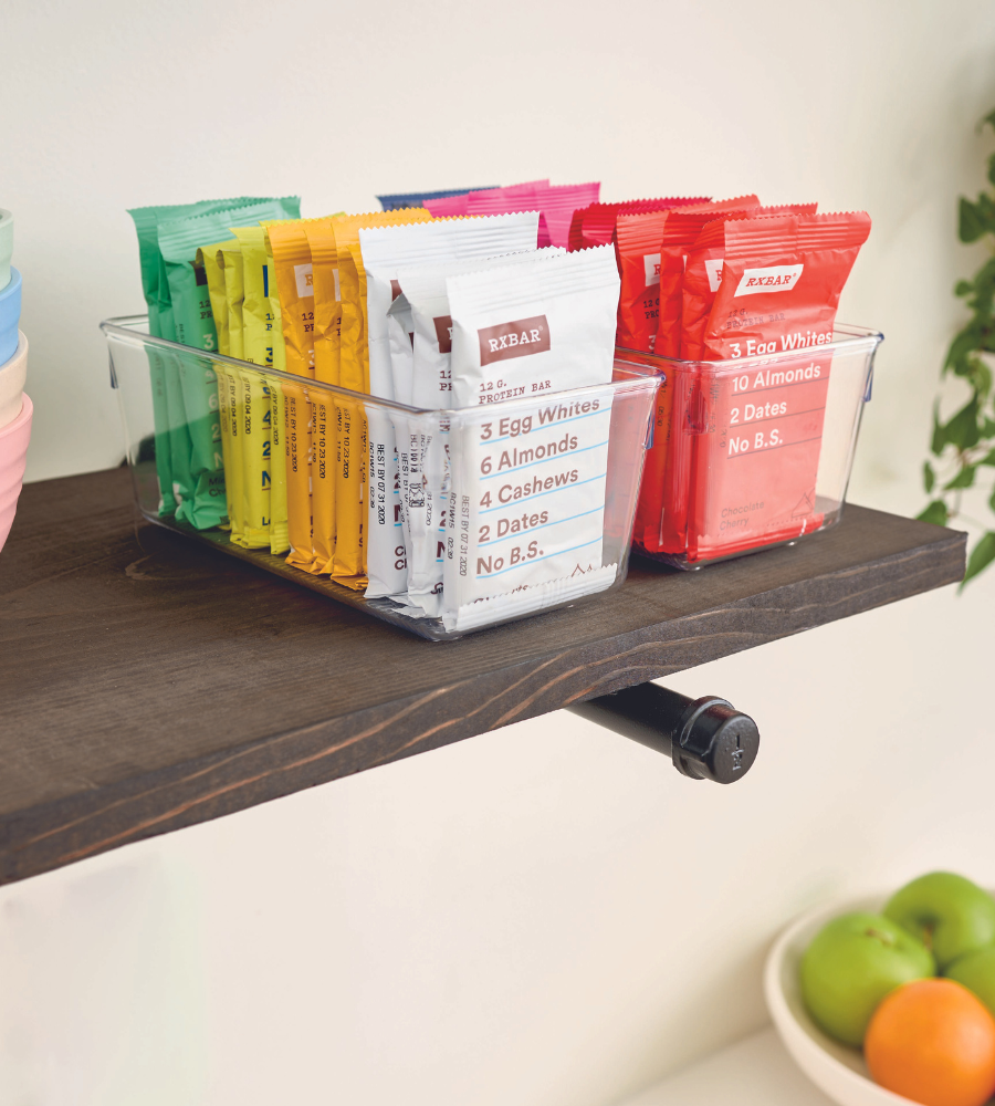 An iDesign LINUS HIGH drawer organizer holds colorful RXBAR protein bars on a dark wood shelf, with a bowl of fruit underneath.