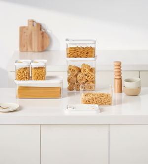 Sul bancone della cucina ci sono dei contenitori impilabili DATEMATE KLAR bianchi con timer di cottura di BNB x VCN pieni di pasta, ciotole impilate, macina sale e pepe in legno, un piatto con una forchetta e taglieri appoggiati al muro.