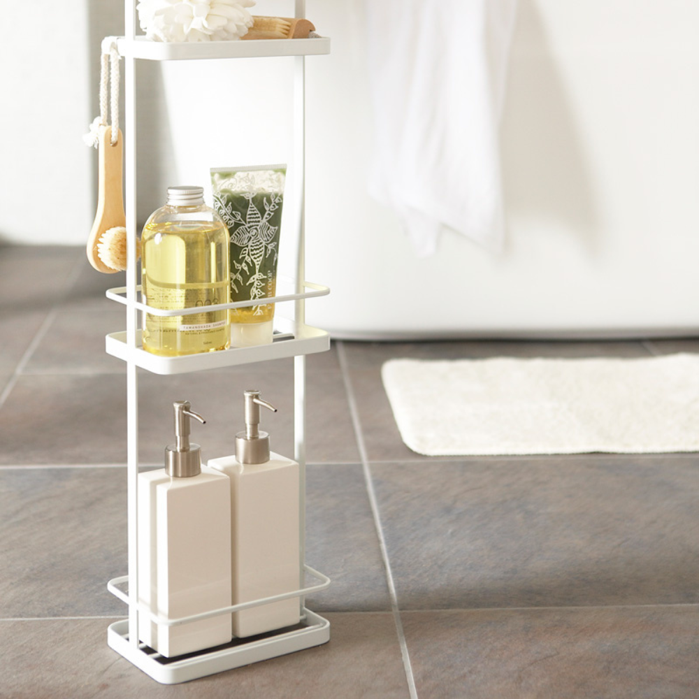 Das YAMAZAKI TOWER Badregal in Weiß ordnet Seife, Lotion, Tiegel, Bürste und Schwamm auf einem grauen Fliesenboden neben weißer Badewanne und Badteppich und bietet extra Stauraum.