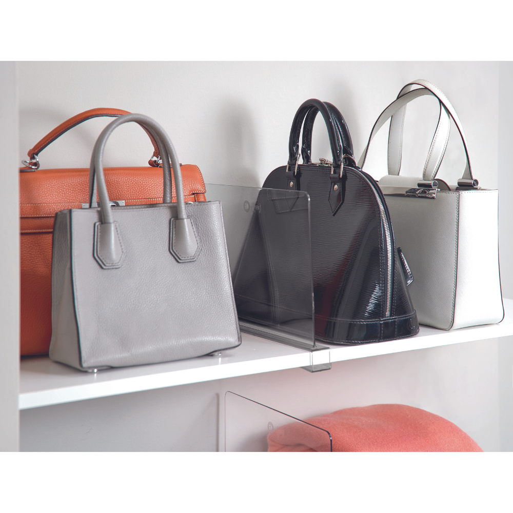 Drei Handtaschen - orange, hellgrau und schwarz glänzend - werden von iDesign HOME EDIT - Regaltrenner KLAR auf einem weißen Regal getrennt. Ein gefalteter pfirsichfarbener Gegenstand liegt auf dem unteren Regal.