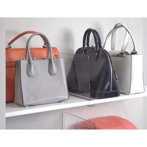 Drei Handtaschen - orange, hellgrau und schwarz glänzend - werden von iDesign HOME EDIT - Regaltrenner KLAR auf einem weißen Regal getrennt. Ein gefalteter pfirsichfarbener Gegenstand liegt auf dem unteren Regal.