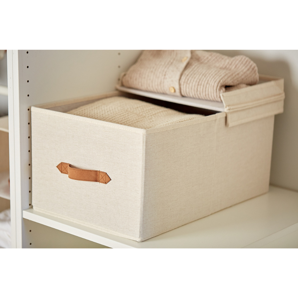 Die PREMIUM Aufbewahrungsbox mit Klappdeckel - Beige von Love it Store it mit braunem Griff steht auf einem weißen Regal, dessen Deckel teilweise geöffnet ist und in dem sich ordentlich gefaltete cremefarbene Pullover befinden.