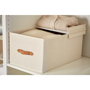 Die PREMIUM Aufbewahrungsbox mit Klappdeckel - Beige von Love it Store it mit braunem Griff steht auf einem weißen Regal, dessen Deckel teilweise geöffnet ist und in dem sich ordentlich gefaltete cremefarbene Pullover befinden.