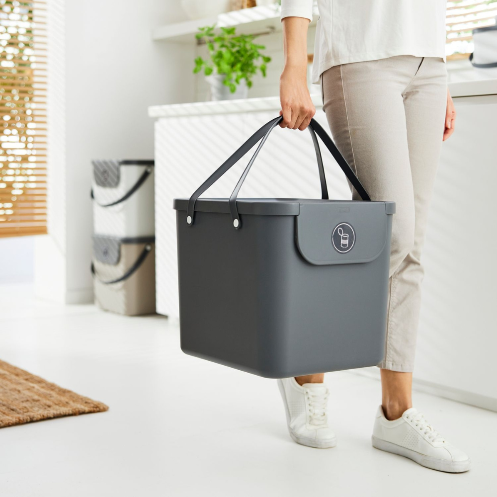 Eine leicht bekleidete Person trägt einen großen, anthrazitfarbenen Rotho 3er Set - ALBULA Recycling Müllsystem 40L mit schwarzen Griffen in einer hellen Küche mit weißen Schränken und Grünpflanzen, die eine effiziente Mülltrennung zeigt.