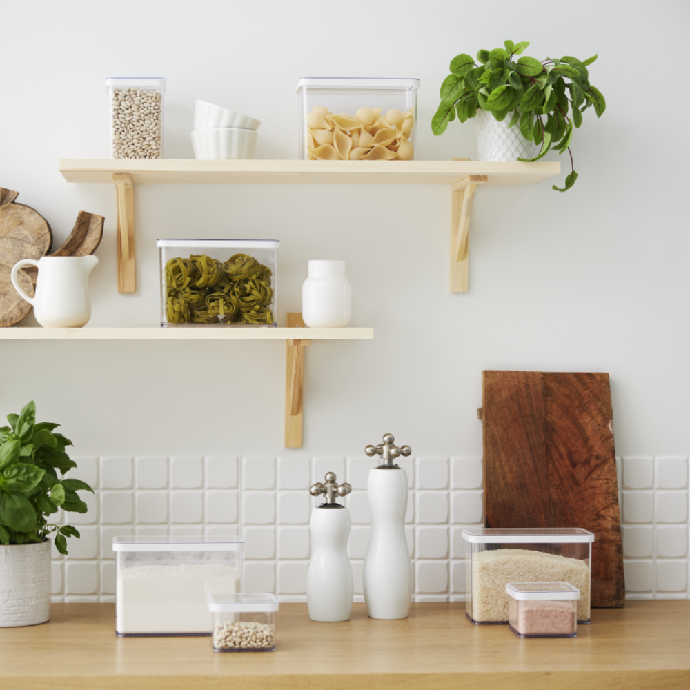 Auf einer modernen Küchenarbeitsplatte ist das Rotho Vorratsdosen-Set LOFT (6-teilig) mit Nudeln, Körnern und Bohnen übersichtlich angeordnet. Darüber befinden sich Holzregale mit weiteren Behältern und Geschirr. Weiße Fliesen und helle Holzakzente vervollständigen den frischen Look.