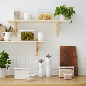 Auf einer modernen Küchenarbeitsplatte ist das Rotho Vorratsdosen-Set LOFT (6-teilig) mit Nudeln, Körnern und Bohnen übersichtlich angeordnet. Darüber befinden sich Holzregale mit weiteren Behältern und Geschirr. Weiße Fliesen und helle Holzakzente vervollständigen den frischen Look.