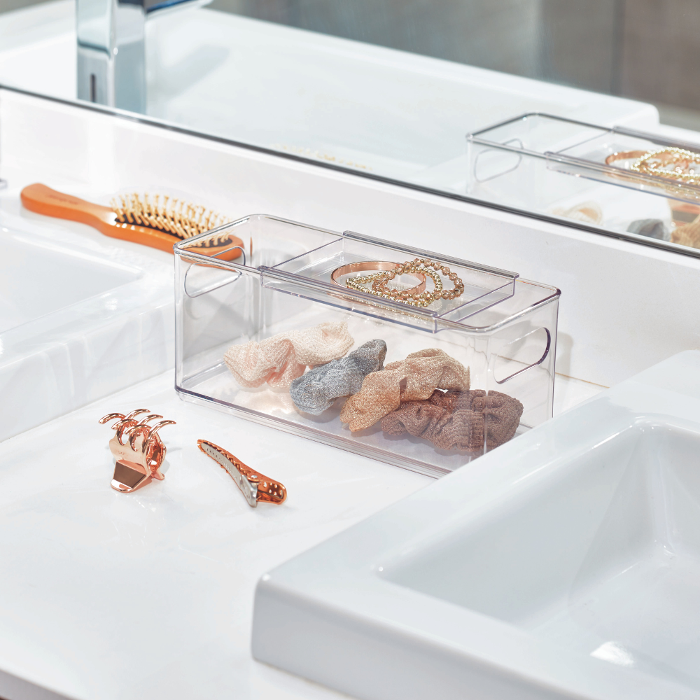 Ein iDesign HOME EDIT- Aufbewahrungsbehälter - Accessory steht auf einem weißen Badezimmertisch, in dem Haargummis und Schmuck ordentlich aufbewahrt werden. In der Nähe befinden sich eine goldene Haarspange, ein Kamm, zwei Waschbecken und ein Spiegel, der den organisierten Raum reflektiert.