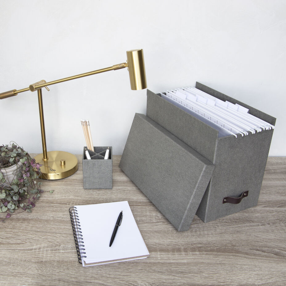 Ein moderner Schreibtisch mit einer goldenen Lampe, einer kleinen Pflanze, einem Stifthalter, einem aufgeschlagenen Notizbuch und einer Bigso JOHAN Hängeregister-Box aus grauem Segeltuch für organisierte A4-Dokumente auf einer Holzoberfläche.