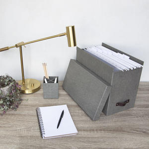 Ein moderner Schreibtisch mit einer goldenen Lampe, einer kleinen Pflanze, einem Stifthalter, einem aufgeschlagenen Notizbuch und einer Bigso JOHAN Hängeregister-Box aus grauem Segeltuch für organisierte A4-Dokumente auf einer Holzoberfläche.