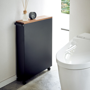 Der TOWER Aufbewahrungswagen mit Griff von YAMAZAKI in Schwarz steht mit Holzplatte und Rollen neben einem modernen weißen WC im minimalistischen Bad und trägt eine Uhr sowie einen Reed Diffuser.