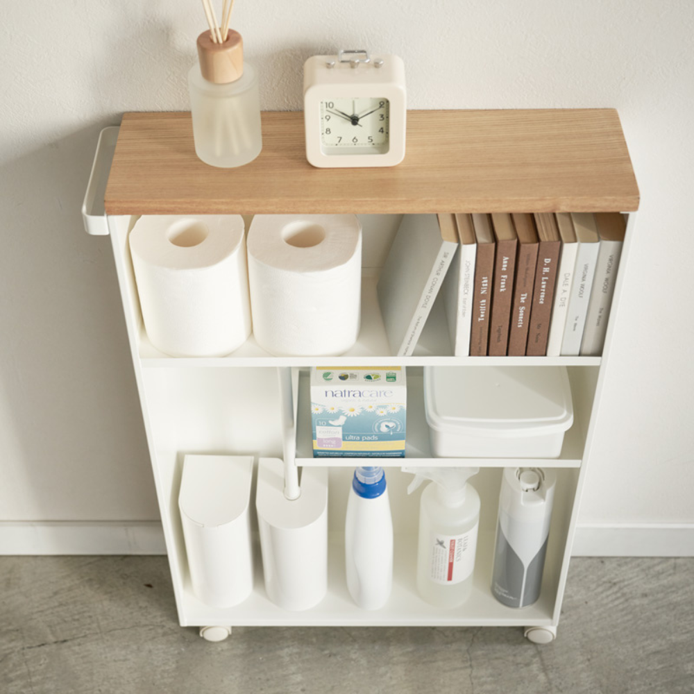 Der YAMAZAKI TOWER Aufbewahrungswagen mit Griff in Weiss, mit Holzplatte, steht an einer weißen Wand auf grauem Boden und bietet zusätzlichen Badezimmer Stauraum für Uhr, Diffusor, Toilettenpapier, Bücher und mehr.