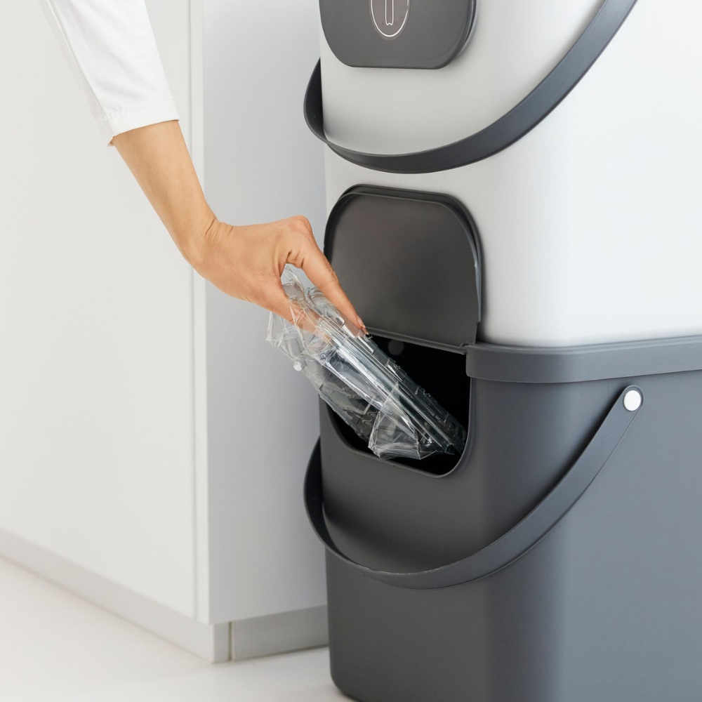 Die Hand einer Person stellt einen durchsichtigen Kunststoffbehälter in einen Rotho ALBULA Recycling Müllsystem 40L - Anthrazit Behälter mit einer seitlichen Öffnung.