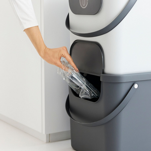 Die Hand einer Person stellt einen durchsichtigen Kunststoffbehälter in einen Rotho ALBULA Recycling Müllsystem 40L - Anthrazit Behälter mit einer seitlichen Öffnung.