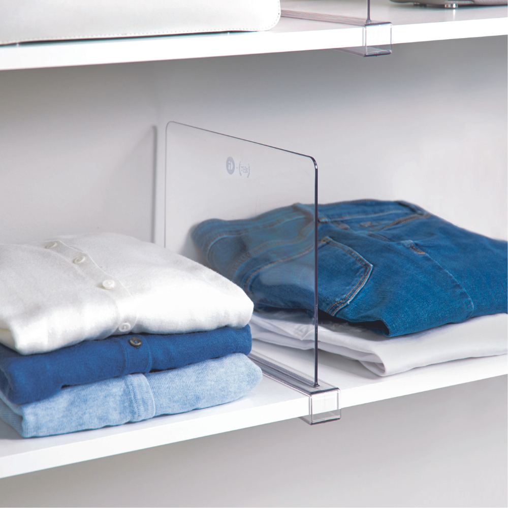 Drei ordentlich gefaltete Stapel weißer, blauer und jeansfarbener Kleidung werden durch den transparenten Regalteiler HOME EDIT - Regaltrenner KLAR von iDesign auf einem weißen Regal getrennt.