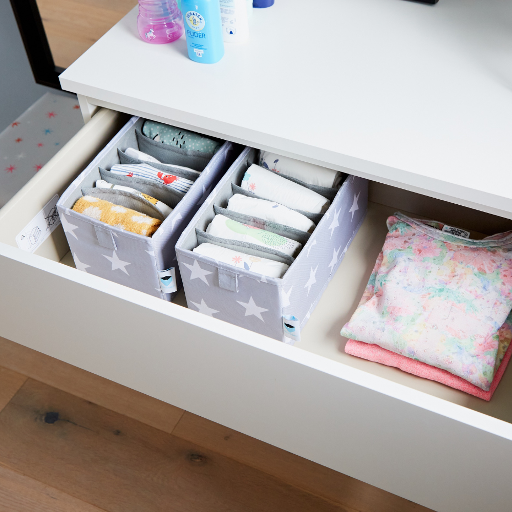 In einer offenen weißen Schublade befinden sich 2 Love it Store it STAR Organizer 6 Fächer schmal Grau Sets mit Babykleidung und Accessoires, daneben pastellfarbene Babykleidung und darüber ordentlich angeordnete Toilettenartikel - ideal fürs Kinderzimmer.