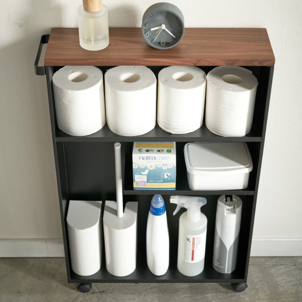 Der TOWER Aufbewahrungswagen mit Griff von YAMAZAKI in Schwarz bietet auf drei Ablagen Platz für fünf Toilettenpapierrollen, eine kleine Uhr, Flaschen, eine Box Binden, einen Behälter und Reinigungszubehör an einer weißen Wand.