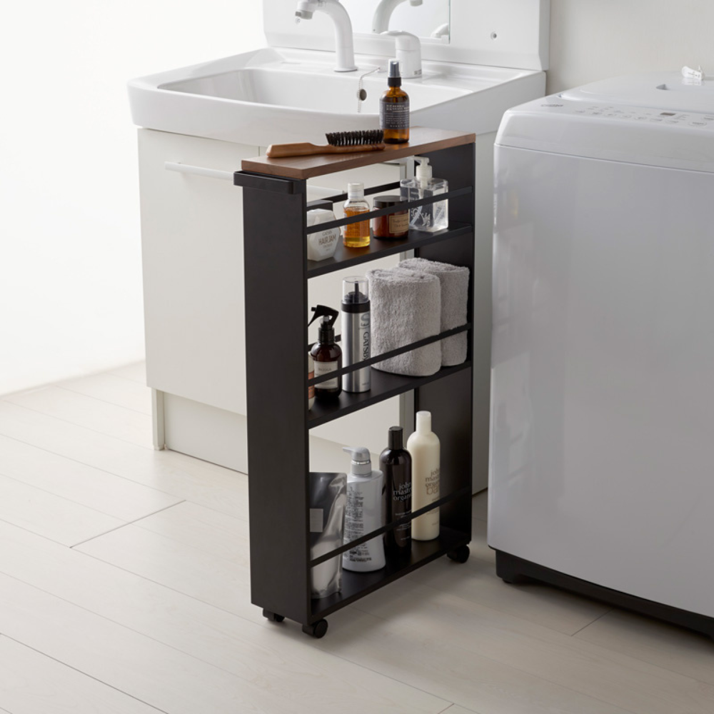 Der YAMAZAKI TOWER Küchenwagen mit Griff in Schwarz, bestückt mit Toilettenartikeln, Handtüchern und Flaschen, steht zwischen weißem Waschbecken und Waschmaschine im minimalistischen Bad und schafft smarten Stauraum.