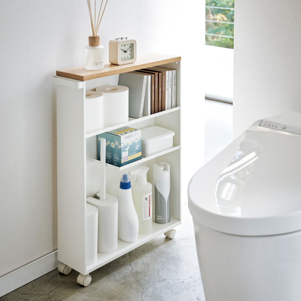 Der YAMAZAKI TOWER Aufbewahrungswagen mit Griff in Weiß, der neben einer modernen weißen Toilette platziert ist, verstaut ordentlich Reinigungsmittel, Toilettenpapier und Bücher. Eine Uhr, ein Diffusor und ein Tablett auf der Oberseite fügen dem hellen Badezimmer einen cleveren Stauraum hinzu.