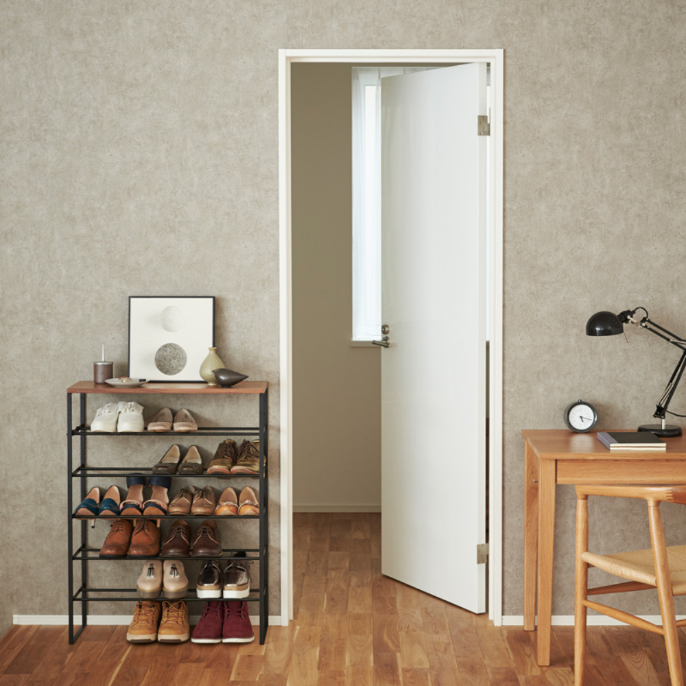 In einem aufgeräumten Raum steht das YAMAZAKI TOWER Schuhregal Holz - Schwarz, ein platzsparendes 6-stöckiges Schuhregal mit Schuhen, gerahmter Kunst und Dekoration. In der Nähe befindet sich ein hölzerner Schreibtisch mit Lampe, Uhr, Stuhl und einer weißen Tür, die ein helles Fenster freigibt.