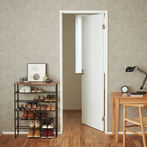 In einem aufgeräumten Raum steht das YAMAZAKI TOWER Schuhregal Holz - Schwarz, ein platzsparendes 6-stöckiges Schuhregal mit Schuhen, gerahmter Kunst und Dekoration. In der Nähe befindet sich ein hölzerner Schreibtisch mit Lampe, Uhr, Stuhl und einer weißen Tür, die ein helles Fenster freigibt.