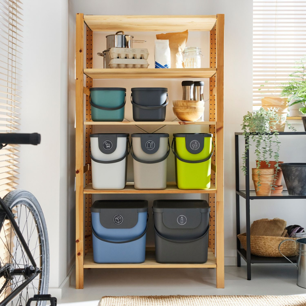 In einem Holzregal stehen bunte Behälter, Küchenutensilien und das Rotho 3er-Set ALBULA Recycling Müllsystem 25L in Anthrazit. Mit Pflanzen und einem Fahrrad in der Nähe schafft das Sonnenlicht durch die Jalousien einen gemütlichen Raum, der die Mülltrennung unterstützt.