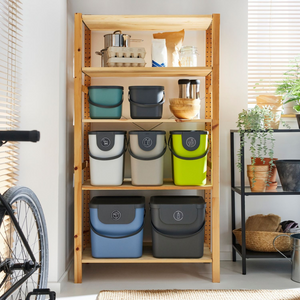 In einem Holzregal stehen bunte Behälter, Küchenutensilien und das Rotho 3er-Set ALBULA Recycling Müllsystem 25L in Anthrazit. Mit Pflanzen und einem Fahrrad in der Nähe schafft das Sonnenlicht durch die Jalousien einen gemütlichen Raum, der die Mülltrennung unterstützt.