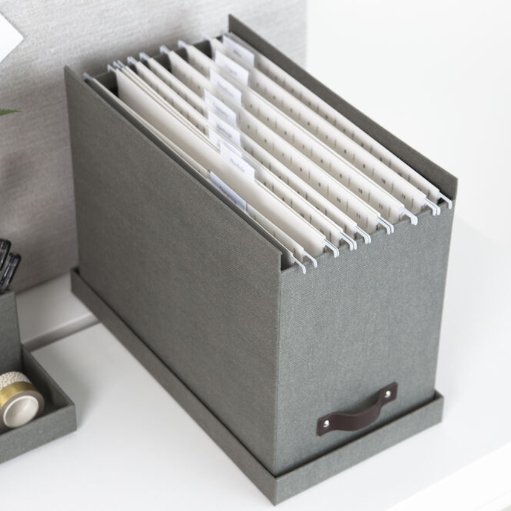 Die Bigso JOHAN Hängeregister-Box in Grau Canvas steht auf einer weißen Fläche und ist mit beschrifteten A4-Hängemappen gefüllt. Die Box verfügt über einen Griff an der Vorderseite, und Büroartikel in der Nähe unterstützen eine effiziente Dokumentenorganisation.