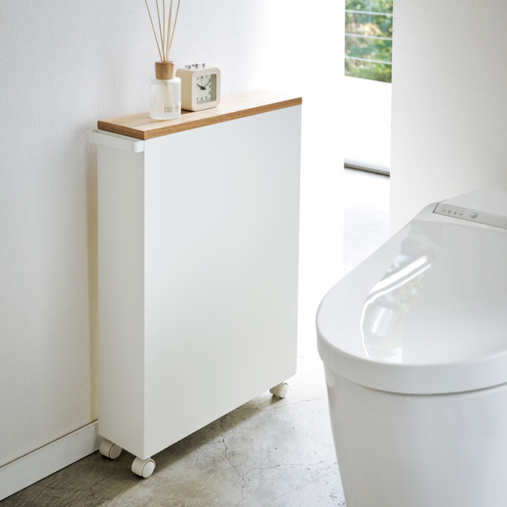 Der YAMAZAKI TOWER Aufbewahrungswagen mit Griff in Weiß, mit Holzablage und Rollen, steht neben einer modernen weißen Toilette und bietet stilvollen Badezimmer Stauraum. Obenauf stehen eine kleine Uhr, eine Raumduft und eine Flasche im Sonnenlicht.