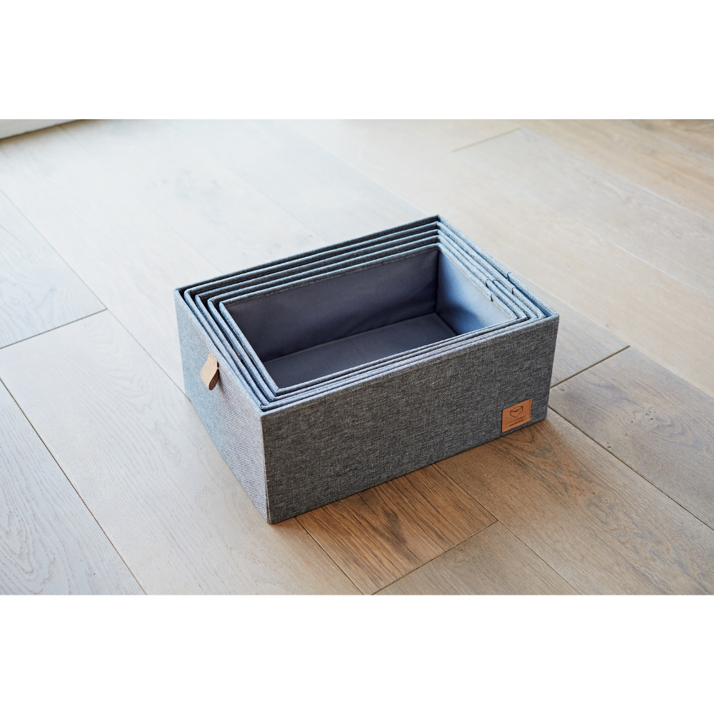 Fünf rechteckige, graue Stoff-Aufbewahrungsboxen aus dem PREMIUM Aufbewahrungsboxen mit Deckel Grau - Set 5 Stk. von Love it Store it stehen ineinander verschachtelt auf einem Holzboden. Die größte Box hat einen kleinen braunen Lederanhänger-perfekt für mehr Ordnung Zuhause.