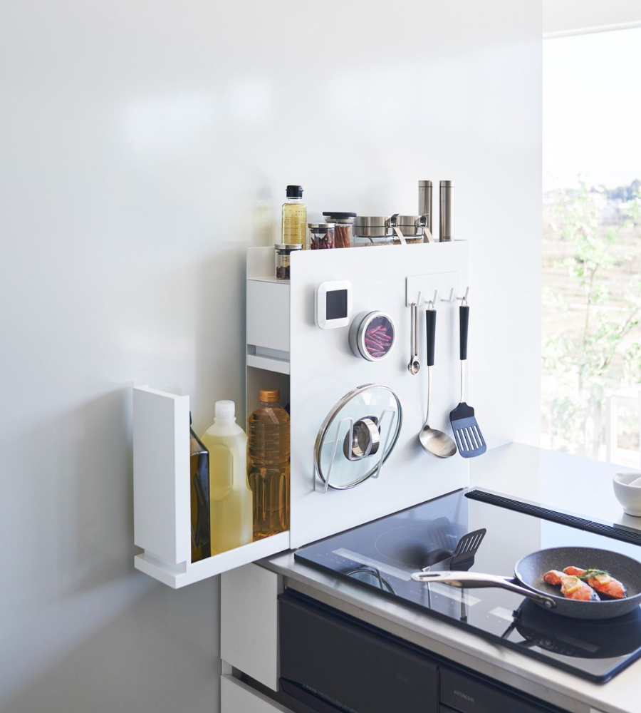 Eine moderne, minimalistische Küchenecke präsentiert das YAMAZAKI TOWER Gewürzregal 2-stöckig - Weiss organisiert Utensilien, Gewürze, Flaschen und einen Pfannendeckel über einem eleganten Induktionsherd, durch dessen Fenster das Sonnenlicht strömt.