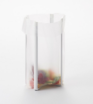 Eine durchsichtige Plastiktüte hängt geöffnet an einem YAMAZAKI TOWER Mehrzweckständer Küche - Weiss, mit bunten Essensresten am Boden, vor einem schlichten weißen Hintergrund.