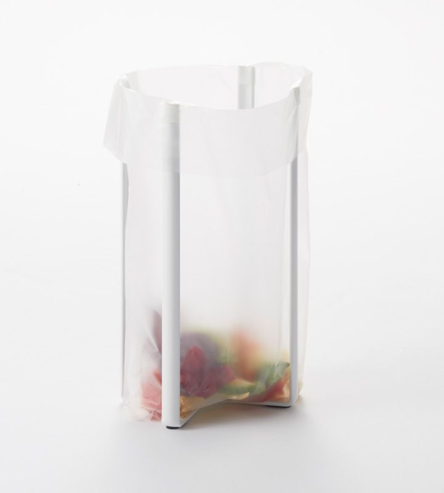 Eine durchsichtige Plastiktüte hängt geöffnet an einem YAMAZAKI TOWER Mehrzweckständer Küche - Weiss, mit bunten Essensresten am Boden, vor einem schlichten weißen Hintergrund.