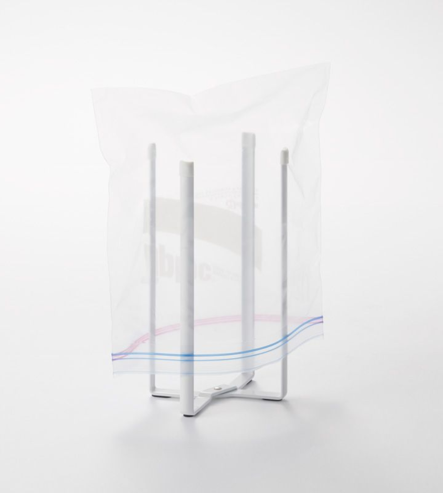 Der YAMAZAKI TOWER Mehrzweckständer Küche - Weiss hält einen durchsichtigen Plastik-Ziplock-Beutel offen. Dieser faltbare, vierzackige Küchenständer vereinfacht die Arbeit und wird vor einem schlichten weißen Hintergrund gezeigt.