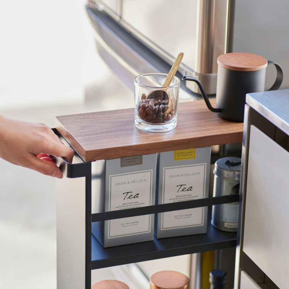 Eine Hand zieht den YAMAZAKI TOWER Küchenwagen mit Griff in Schwarz, auf dessen Holzplatte Kaffeebohnen, ein Löffel und eine schwarze Teekanne stehen. Teedosen auf der unteren Ablage sorgen für praktischen Stauraum.
