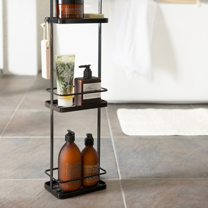 Das YAMAZAKI TOWER Badregal in Schwarz bietet cleveren Stauraum für Seifenspender, Zahnputzglas, Schwamm und Badutensilien auf Fliesenboden neben weißer Badematte und Wanne.